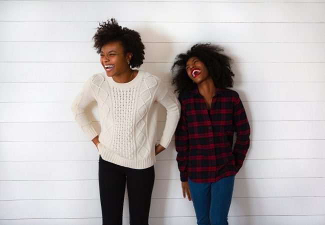Two dark-haired young women stand against a white wall smiling and laughing while wearing long-sleeved sweaters