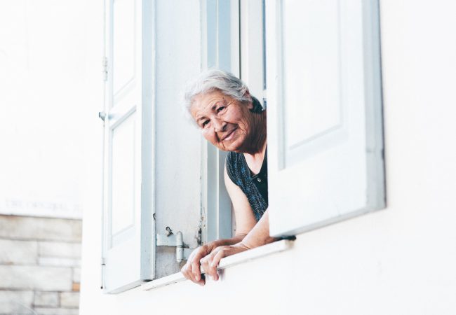 Elderly woman in need of oral care looks out the window of a white building with white shutters